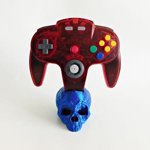 May include: A red Nintendo 64 controller sits on top of a blue skull-shaped stand. The controller has the words "HOME" and "RESET" printed on it.