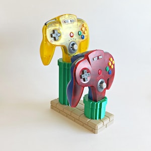 May include: A yellow translucent video game controller and a red translucent video game controller mounted on a green pipe-shaped stand with a tan brick-patterned base. The controllers feature a central analog stick, a directional pad, and buttons.