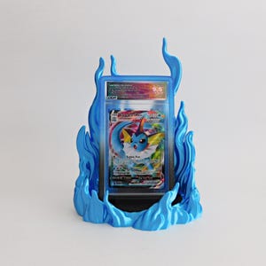 May include: A graded Vaporeon VMAX trading card encased in a clear plastic slab, displayed within a bright blue, flame-shaped stand. The card features a blue creature illustration, the text Vaporeon VMAX, and a DGA 9.5 grade label at the top.