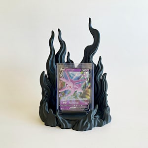 May include: A matte black display stand shaped like stylized flames, holding a clear plastic protective case containing a purple Espeon V trading card. The card features the character Espeon, the text Espeon V, and a health value of 200.