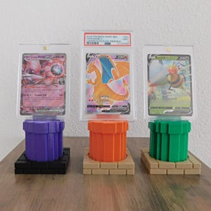 May include: Three graded Pokémon trading cards displayed on colorful, 3D-printed stands. The cards are encased in clear protective holders. The stands are designed to resemble video game elements, with a purple, orange, and green pipe design.