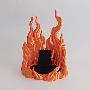 May include: A display stand featuring a sculpted orange flame design surrounding a central black, trapezoidal jewelry holder. The vibrant orange flames rise in varying heights around the dark, solid base, creating a structured display piece.