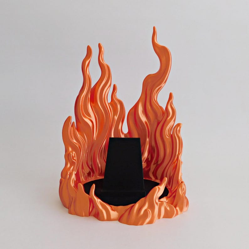 May include: A display stand featuring a sculpted orange flame design surrounding a central black, trapezoidal jewelry holder. The vibrant orange flames rise in varying heights around the dark, solid base, creating a structured display piece.