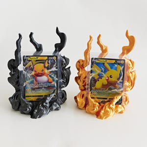 May include: Two trading card display stands shaped like stylized flames. One stand is matte black and holds a Raichu V card, while the other is metallic gold and holds a Pikachu V card. Each card is encased in a clear protective plastic sleeve.