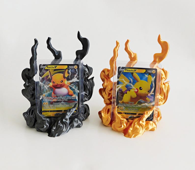May include: Two trading card display stands shaped like stylized flames. One stand is matte black and holds a Raichu V card, while the other is metallic gold and holds a Pikachu V card. Each card is encased in a clear protective plastic sleeve.