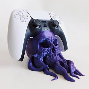 May include: A white and black PlayStation 5 controller rests on a purple skull-shaped stand. The stand has a detailed, textured design with flowing tentacles. The controller is centered above the skull, creating a unique gaming accessory.