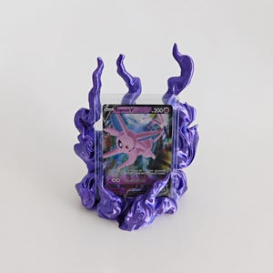 May include: A purple, flame-shaped display stand holding a clear plastic card protector. Inside the protector is a trading card featuring a pink Espeon character with the text Espeon V and 200 HP printed on the top right corner of the card.