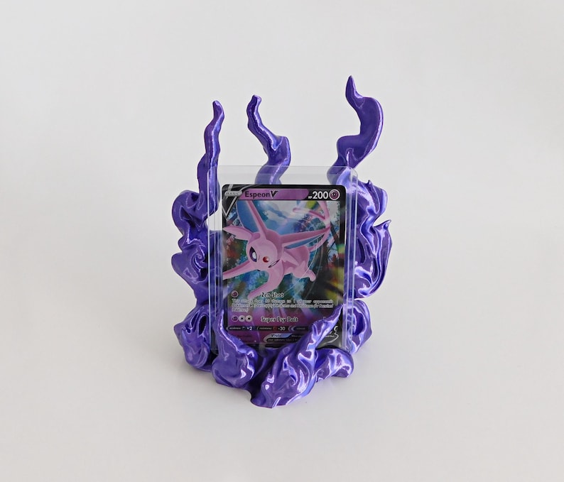 May include: A purple, flame-shaped display stand holding a clear plastic card protector. Inside the protector is a trading card featuring a pink Espeon character with the text Espeon V and 200 HP printed on the top right corner of the card.