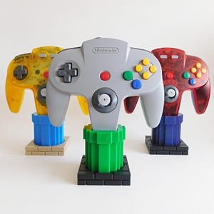 May include: Three Nintendo 64 game controllers displayed on stands. The center controller is solid gray, the left is translucent yellow, and the right is translucent red. Each controller rests on a plastic pipe-shaped base with a square brick-style foundation.