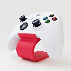 May include: A white Xbox controller is displayed on a red stand. The controller has black buttons and a white and green Xbox logo.