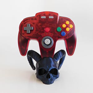 May include: A red translucent N64 controller rests on a dark blue skull-shaped stand with curved horns. The controller has gray, yellow, green, and blue buttons. The stand is a detailed, 3D-printed design.