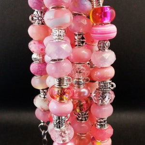 May include: A pink and silver charm bracelet with various beads, including clear, opaque, and patterned beads. Some beads have silver accents and lettering.