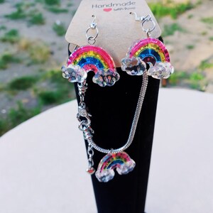 May include: Rainbow charm earrings and bracelet set with silver chains. The charms are made of clear resin with a rainbow glitter design and white clouds. The card behind the jewelry says "Handmade with love."