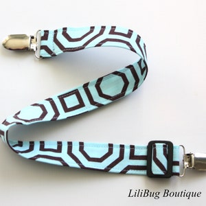 May include: A baby pacifier clip with a geometric pattern in brown and light blue. The clip has two metal clasps and a black plastic slider. The fabric is patterned with brown geometric shapes on a light blue background.