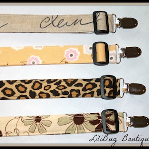 May include: Four fabric straps with metal clips. The straps feature different patterns: beige with black writing, yellow with pink flowers, brown and black leopard print, and beige with green flowers. The straps are all the same length and have a black buckle.