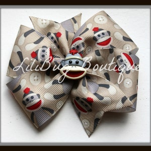 May include: A brown and gray hair bow with a monkey face button in the center. The bow is made of fabric with a pattern of monkeys wearing red hats. The bow is tied in a swirl shape.