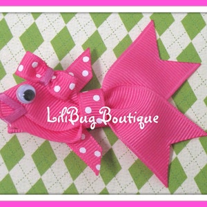 May include: A pink fish-shaped hair bow with white polka dots and a single googly eye. The bow is made of ribbon and has a small loop at the top for attaching to hair.