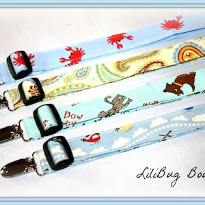 May include: Four colorful fabric straps with metal clips. The straps feature different patterns, including a paisley design, a blue and white cloud pattern, a blue and white pattern with a dog, and a blue pattern with red crabs. The straps are labeled "LiliBug Boutique".
