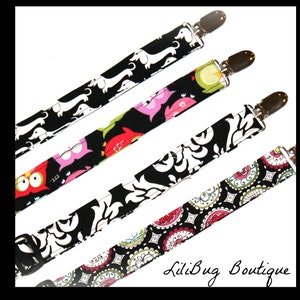 May include: Four colorful fabric baby bib clips with different patterns, including dachshunds, owls, paisley, and a damask design. The clips are silver metal and have a spring-loaded mechanism. The text "LiliBug Boutique" is visible in the bottom right corner of the image.