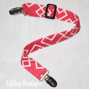 May include: A pink and white patterned fabric strap with two silver clips and a black plastic buckle.  The strap is designed to be used as a pacifier holder or a teether holder.