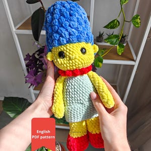 May include: A handmade crochet doll with a blue bouffant hairstyle, yellow skin, and a red necklace. The doll wears a light green dress and red shoes. The doll is a character from a popular animated TV show. The image includes the text "English PDF pattern".