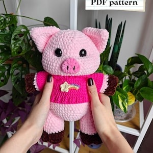 Puede incluir: Un cerdo rosa hecho a mano con ganchillo, con una camisa magenta y pezuñas marrones. El cerdo tiene ojos negros y un pequeño hocico rosa. La camisa presenta un diseño de estrella y arcoíris. Incluye el texto "English PDF pattern".