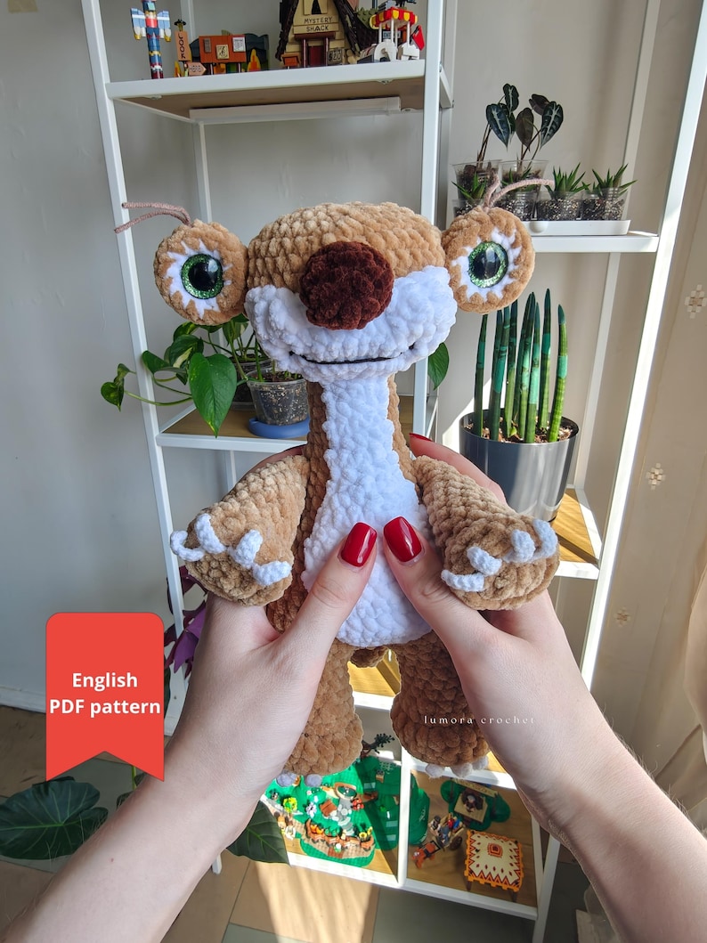 May include: A handmade crochet plush toy in shades of brown, white, and tan, resembling a cartoon character. The toy has large, expressive eyes with green irises, a brown nose, and white claws. The text "English PDF pattern" is visible on a red square.
