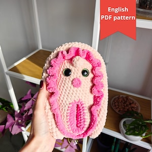 以下が含まれることがあります： 手作りの、ぬいぐるみのような、ピンクとクリーム色の編み人形。人形は、大きな緑色の動く目と、明るいピンク色のテクスチャードアウトラインが特徴です。「English PDF pattern」というテキストが赤いタグに表示されています。
