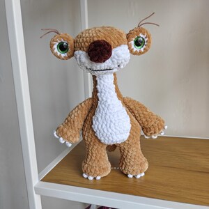 May include: A handmade crochet plush toy of a cartoon character. The plush has a brown body, white chest, and a brown nose. It has large, round eyes with green irises and white accents. The toy is standing on a wooden shelf.