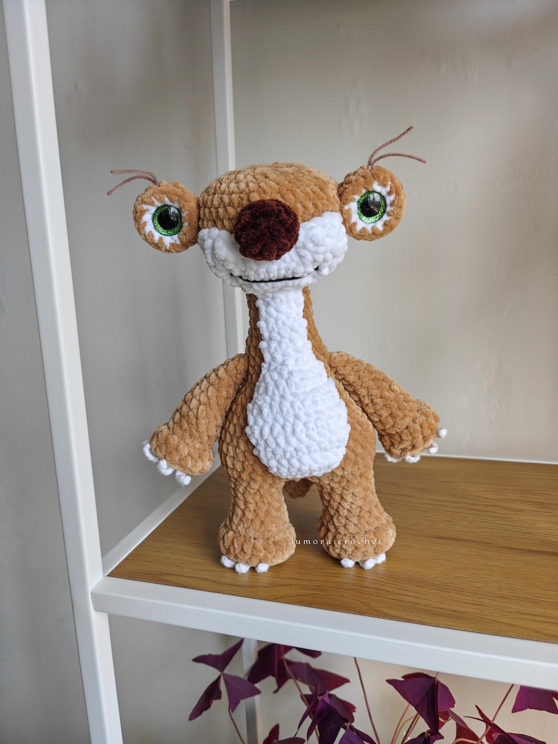 May include: A handmade crochet plush toy of a cartoon character. The plush has a brown body, white chest, and a brown nose. It has large, round eyes with green irises and white accents. The toy is standing on a wooden shelf.