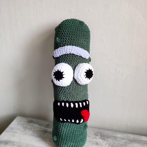 Può includere: Giocattolo di peluche verde lavorato a uncinetto a forma di cetriolo, con una fascia bianca, grandi occhi bianchi con pupille nere, una bocca nera con denti bianchi e una lingua rossa.