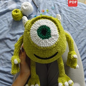 May include: A handmade, green crocheted monster toy with a large, white-rimmed eye and a green center. The toy has small horns, arms, and legs. The image also shows yarn and a PDF icon.
