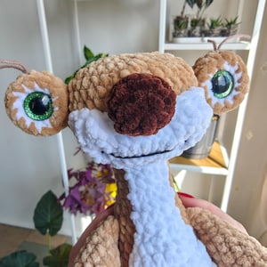 May include: A handmade plush toy of a cartoon sloth character. The sloth has large, round eyes with green glitter accents, a brown nose, and a white and brown body. The toy is made of soft, textured yarn.