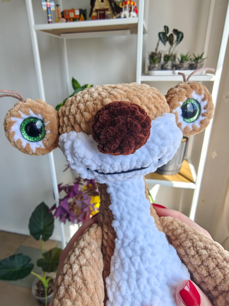 May include: A handmade plush toy of a cartoon sloth character. The sloth has large, round eyes with green glitter accents, a brown nose, and a white and brown body. The toy is made of soft, textured yarn.