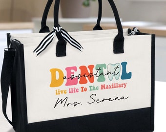 Könnte beinhalten: Weiße Canvas-Tragetasche mit schwarzen Griffen und schwarzem Boden. Die Tasche hat eine bunte Grafik mit der Aufschrift "Dental Assistant" mit einer Zahngrafik und dem Text "live life to the maxillary" und "Mrs. Serena".