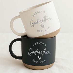 May include: Two stacked ceramic mugs. The top mug is off-white with a brown base and a curved handle. The bottom mug is black with a brown base and a curved handle. Both mugs have the text "Katlyn's Godmother" and "EST. 2025".