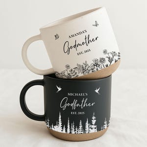 May include: Two ceramic mugs stacked on top of each other. The top mug is off-white with the words "Amanda's Godmother EST. 2025" and floral and butterfly designs. The bottom mug is black with "Michael's Godfather EST. 2025" and a forest design.