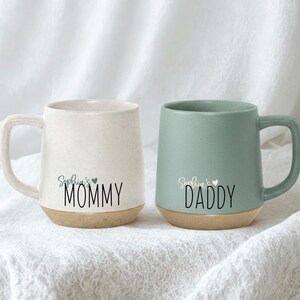 May include: Two ceramic mugs, one cream and one sage green, with brown bases. The cream mug says "Sophia's MOMMY" and the green mug says "Sophia's DADDY" in black script. Both mugs have a small blue heart.