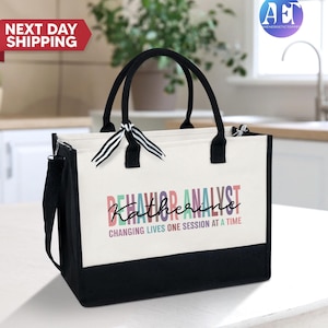 May include: A white canvas tote bag with black handles and a black bottom. The bag has a colorful graphic that says "Behavior Analyst Katherine" and "Changing Lives One Session at a Time".