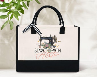 May include: A white canvas tote bag with black handles and a black bottom. The bag has a design of a vintage sewing machine with flowers and the text "SEWCIOPATH" and "Alister" in red.