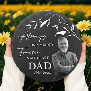 May include: A circular black slate memorial plaque. The text reads "Always on my mind, Forever in my heart, Dad 1942-2025." A black and white photo of a man is on the right. White floral design accents the text.
