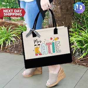 May include: A white canvas tote bag with black handles and a black bottom. The bag has a design of the word "Katie" in colorful letters with school supplies and a red apple. The text "Mrs." is above the name.