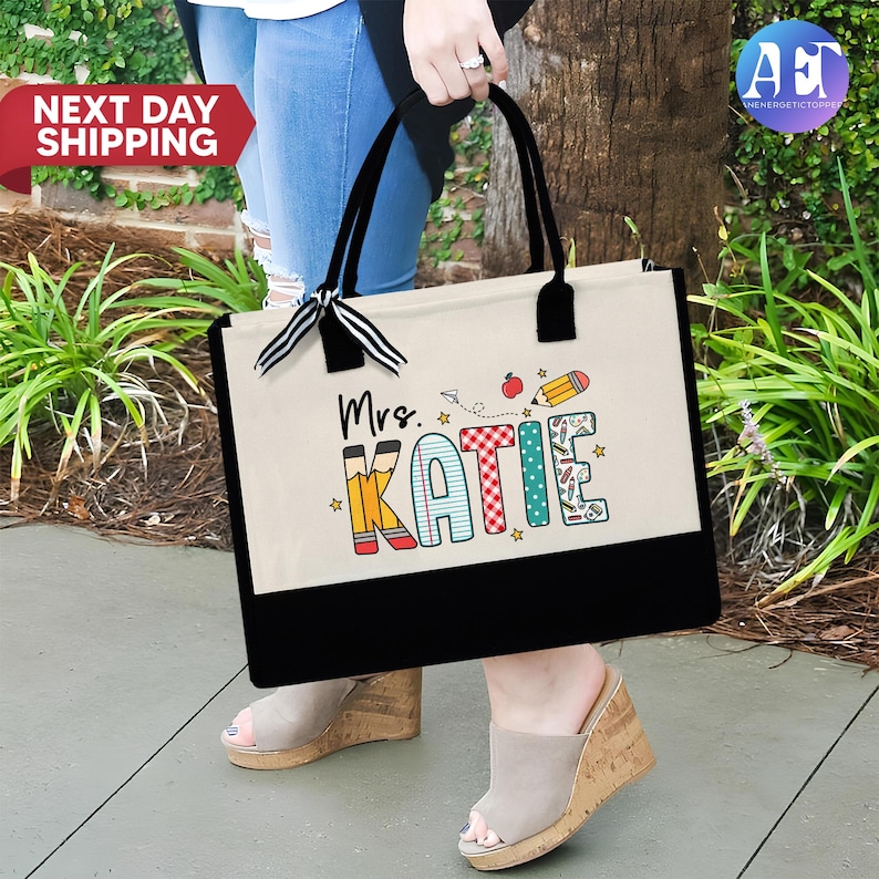 May include: A white canvas tote bag with black handles and a black bottom. The bag has a design of the word "Katie" in colorful letters with school supplies and a red apple. The text "Mrs." is above the name.