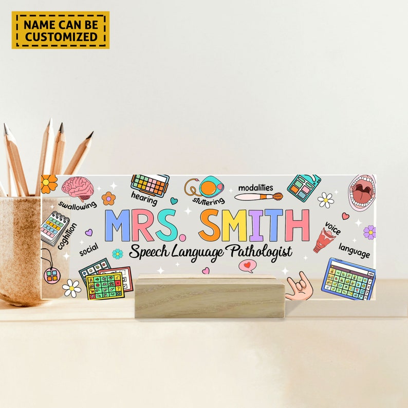 May include: A clear acrylic sign with a wooden stand, featuring colorful illustrations of speech therapy related icons and the text "MRS. SMITH Speech Language Pathologist".