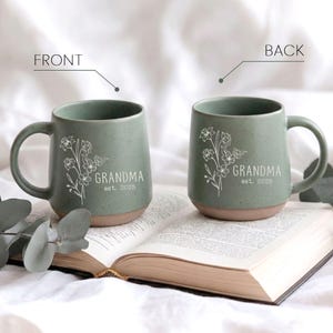 May include: Two sage green ceramic mugs with a floral design and the word "GRANDMA est. 2025" printed in white. The mugs have a rounded shape with a brown base and a curved handle. They are sitting on an open book.