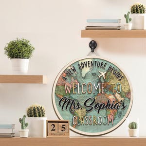 May include: Round wooden sign with a world map design and the text "A New Adventure Begins Welcome to Mrs. Sophia's Classroom". The sign includes a small airplane graphic and is hanging from a black hook. A wooden calendar shows the date September 25.