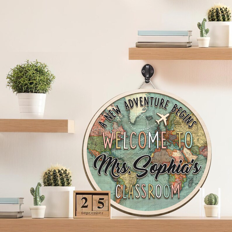 May include: Round wooden sign with a world map design and the text "A New Adventure Begins Welcome to Mrs. Sophia's Classroom". The sign includes a small airplane graphic and is hanging from a black hook. A wooden calendar shows the date September 25.