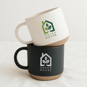 May include: Two ceramic mugs stacked on top of each other. The top mug is white with a green and white logo that says "GREEN HOUSE". The bottom mug is black with a white logo that says "GREEN HOUSE". Both mugs have a tan base.