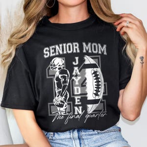 May include: Black t-shirt with a football-themed design. The shirt features the words "SENIOR MOM" above a football and a football player graphic. The name "JAYDEN" is printed on the football. The text "The final quarter" is printed below the football.