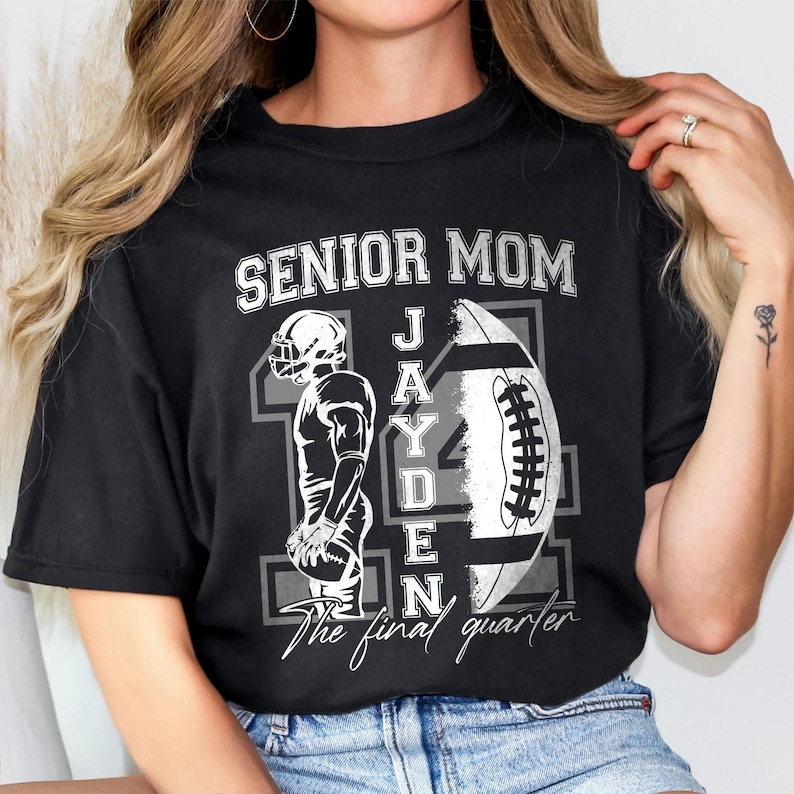 May include: Black t-shirt with a football-themed design. The shirt features the words "SENIOR MOM" above a football and a football player graphic. The name "JAYDEN" is printed on the football. The text "The final quarter" is printed below the football.